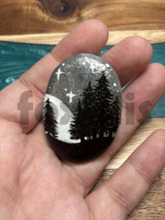 A photo of a rock in a hand. The rock is painted with a moon, stars, and some trees.