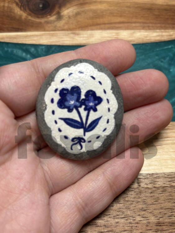 A photo of a rock in a hand. The rock is painted with a white scalloped circle with a couple of vintage-looking flowers in the center.
