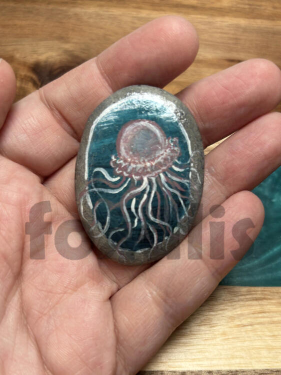 A photo of a rock painted with a blue ombre background with a pink jellyfish painted over it.